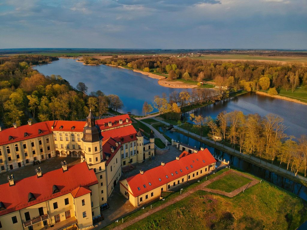 Drone view scenery of beige Nesvizh Castle with brown tiled roofs located on green riverside in Belarus